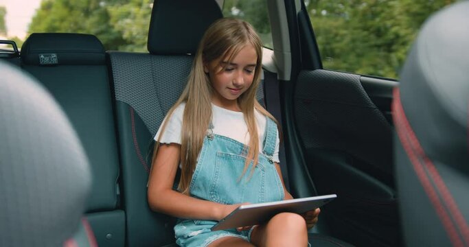 Attaractive little girl passenger in car reading digital tablet and watching cartoons online. Child networking with tablet computer while travelling by car on summer vacation.