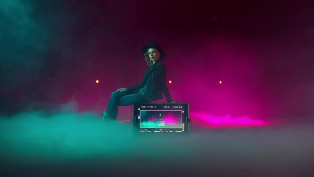 Female model, actress filming in a studio filled with smoke and neon lights background. Young woman in a hat sitting on a playback in the middle of the room.