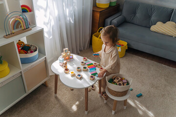 A little girl playing with colorful wooden blocks on the table in playroom. Educational game for toddler in modern nursery. The kid builds from wooden rainbow stacking blocks and learning colors.