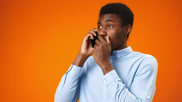 Worried young african man talking on the phone recieving bad news against orange background