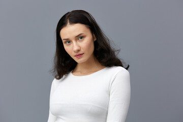 a sweet, pretty woman stands on a gray background in a white T-shirt, hid her hands behind her back, slightly frowning her eyebrows, looking around, turning her body slightly to the side