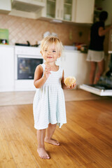 Little girl with a bun in her hand stands on a wooden floor in the kitchen. High quality photo