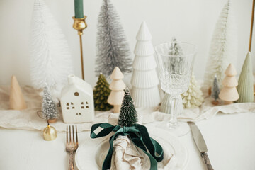 Christmas table setting. Stylish napkin with ribbon and bell on plate, vintage cutlery, modern festive christmas trees and houses on white rustic table. Holiday arrangement of table