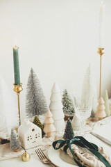 Stylish Christmas table setting. Festive napkin with ribbon and bell on plate, vintage cutlery, wineglass, modern christmas trees and houses on white rustic table. Holiday brunch