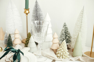 Stylish Christmas table setting. Festive napkin with ribbon and bell on plate, vintage cutlery, wineglass, modern christmas trees and houses on white rustic table. Holiday brunch