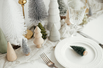 Modern Christmas table setting. Stylish little christmas tree on plate, vintage cutlery, glasses, festive fir and houses on white rustic table. Holiday arrangement of table