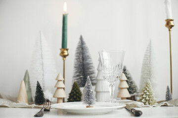 Modern Christmas table setting. Stylish little christmas tree on plate, vintage cutlery, glasses, festive fir and houses on white rustic table. Holiday arrangement of table