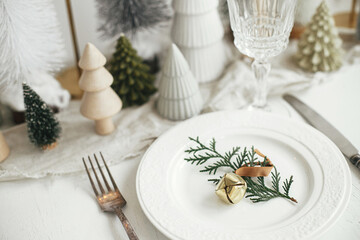 Stylish Christmas table setting. Cedar branch with bell on plate, vintage cutlery, wineglass, modern christmas trees and houses on white rustic table. Holiday brunch, new year celebration