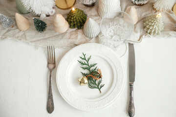 Fototapeta na wymiar Stylish Christmas table setting. Cedar branch with bell on plate, vintage cutlery, wineglass, modern christmas trees and houses on white rustic table. Holiday brunch, top view