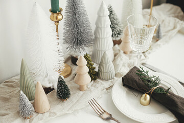 Stylish Christmas table setting. Cotton napkin with bell and fir on plate, vintage cutlery, wineglass, modern christmas trees and candle on white rustic table. Holiday brunch