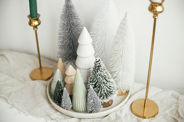 Stylish christmas little trees on plate, linen runner, golden candles on rustic table. Modern white artificial fir trees decoration in room. Holiday arrangement of table, christmas setting