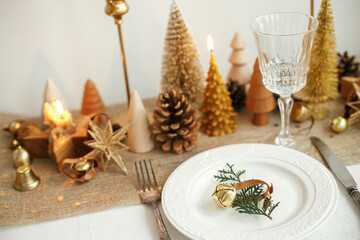 Fototapeta na wymiar Stylish Christmas table setting. Fir branch with bell on plate, vintage cutlery, wineglass, modern golden christmas trees and candles on rustic table. Holiday brunch, new year celebration