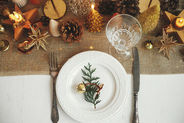 Stylish Christmas table setting. Fir branch with bell on plate, vintage cutlery, wineglass, modern golden christmas trees and candles on  rustic table. Holiday brunch, new year celebration, top view