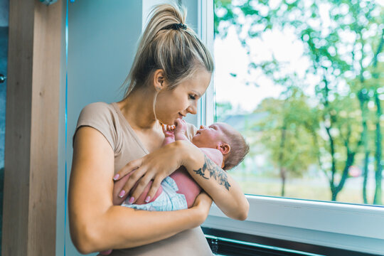 Millennial caucasian blonde mother with hair tied into ponytail holding and smiling at her beautiful first-born son while standing by the window. Blurred trees. High quality photo