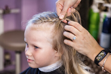 Close up barber woman make fashionable braids hairstyle for cute little blond girl child in modern barbershop, hair salon. Hairdresser makes hairdo for baby in barber shop. Concept hairstyle, beauty