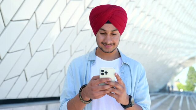 Portrait of cheerful young Indian guy sending message on mobile phone outdoors. Happy man in national headdress using smartphone, enjoying chatting online, messaging in social networks, front view