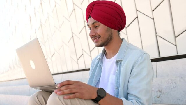 Cheerful indian male student greeting participants of virtual meeting, man in national headdress sits with laptop and waving hello into webcam, has video conference with classmates sitting outdoors