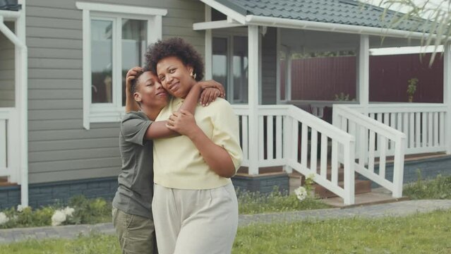 Medium portrait of delighted African American woman and her loving 12 year old son posing for camera standing outside their house in summer