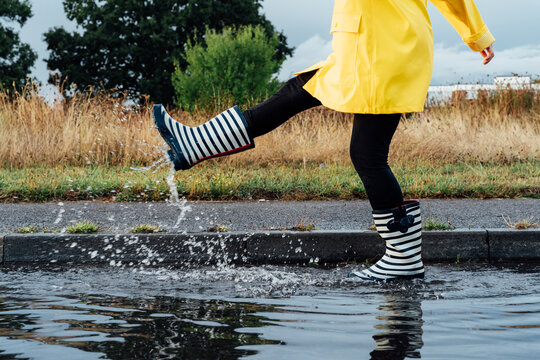 Woman having fun on the street after the rain. Cropped woman wearing rain rubber boots and yellow raincoat walking into puddle with water splash and drops. Fall weather. Selective focus