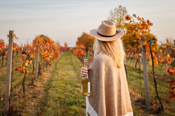 Female vintner enjoying white wine in her vineyard. Woman with poncho and hat holding wine bottle...