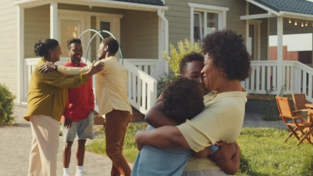 Medium shot of cheerful Black woman greeting and hugging her teenage nephews while getting together for family dinner on green lawn in yard outside house