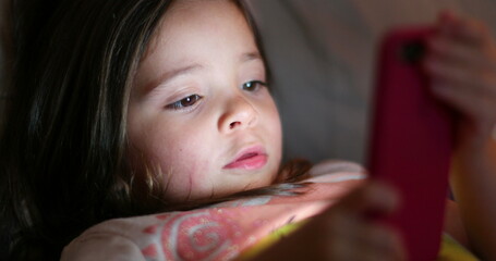 Little girl child using smartphone device. Light glowing on girl face looking at cellphone screen