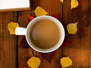 On a wooden table there is a white mug with coffee around the mug lies fallen leaves, next to it lies a book with a brown cover