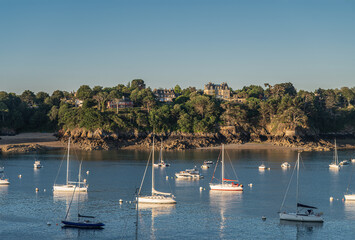 Fototapeta na wymiar St. Malo, Brittany, France - July 8, 2022: Morning light on Brise Moulin Mansion set on green foliage covered rocky shoreline of Rance River mouth under blue sky, White yachts in front