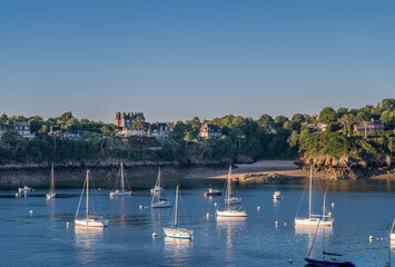 Fototapeta na wymiar St. Malo, Brittany, France - July 8, 2022: Morning light on white yachts on blue Rance river mouth with iconic buildings on green shore of Pointe de la Vicomte, under blue sky.