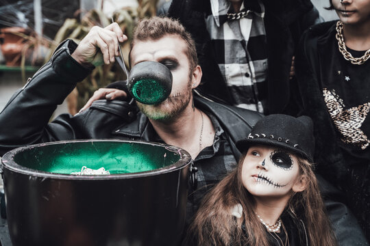 Scary family, mother, father, daughters celebrating halloween. Cauldron of potion.Terrifying black skull half-face makeup,witch costumes,stylish images.Horror,fun at children's party in barn on street