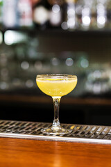 Glass of yellow cocktail decorated with lemon at bar counter background.