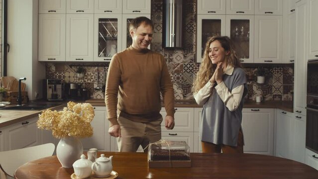 Locked down of man limping and woman walking in kitchen at house at daytime, putting cake on dining table, smiling