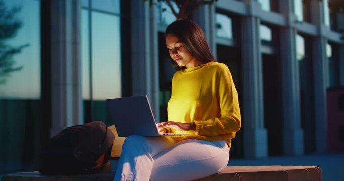 Beautiful young elegant woman sitting on bench outdoor in evening and working on laptop. Pretty Caucasian stylish businesswoman resting at street late and using computer for work or chatting.
