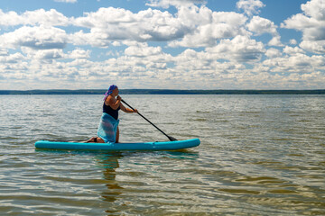 Fototapeta na wymiar a woman in a closed swimsuit, a pareo and a headdress on her knees on a SUP board with an oar floats on the water.