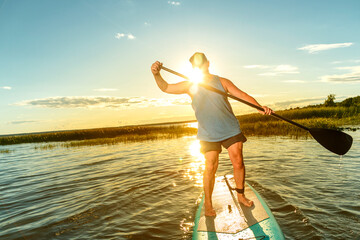 a man on a sup board with an oar floats on the water against the backdrop of the setting sun.