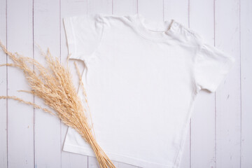 White children's t-shirt mockup for logo, text or design on wooden background with pampas grass top...