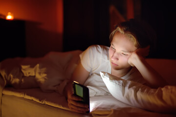 Teenager boy chatting by phone, writing in blog or reading social networks instead sleeping. Young guy laying in bed and watching video clips by smartphone. Overuse and addiction kids from gadgets