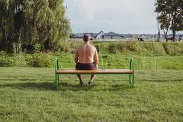 A shirtless man sits alone on a park bench overlooking a field. Solitude. A man with a bare back is sitting on a bench. Field with cows. The man is resting.
