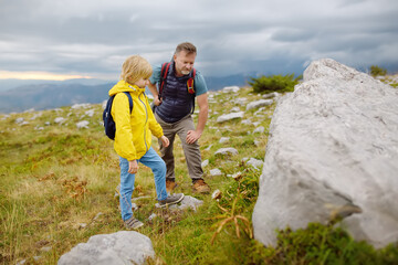 Cute schoolchild and his mature father hiking together on mountain and exploring nature. Child watching a plant. Concepts of adventure, scouting and hiking tourism for kids.