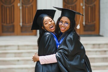 two young ladies in graduation costumes posing at camera at university campus, holding diplomas,...
