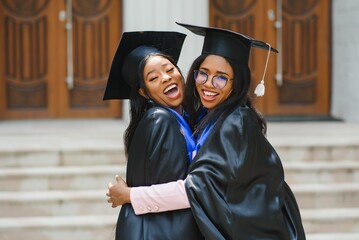 two young ladies in graduation costumes posing at camera at university campus, holding diplomas,...