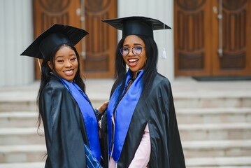 two young ladies in graduation costumes posing at camera at university campus, holding diplomas, laughing and hugging, having happy graduation day, closeup portrait