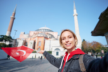 Enjoying vacation in Istanbul. Young traveling woman with national turkish flag taking selfie on view of Hagia Sophia Temple.