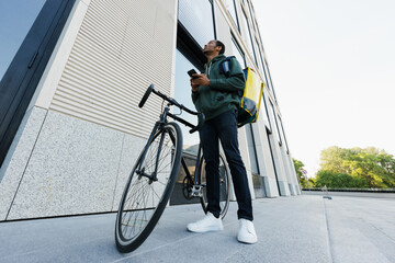 An African-American courier stands next to a bicycle and looks at an office building. The man holds the phone in his hands to check the order.