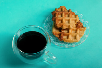 Fluffy waffles. They lie on a saucer. Next to a cup of coffee. Light green background. Shot close-up.