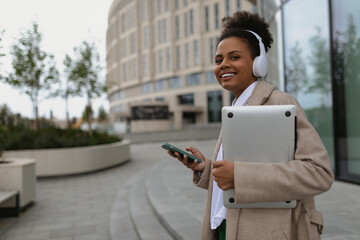 a young American woman with a wide smile walks in headphones with a mobile phone and a laptop against the backdrop of a city building