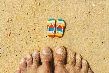 Huge male feet on the beach sand next to miniature beach slippers. Unexpected combination,...