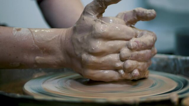 Close-up of potter's hands covered with clay making beautiful vase on throwing wheel in pottery workshop. Creativity and traditional crafts concept.