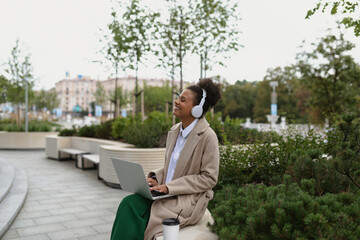 Fototapeta premium a young black student with a laptop sitting on a bench listening to music in headphones with pleasure closing her eyes