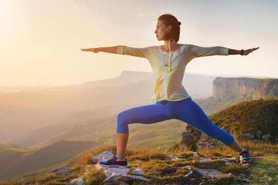 yoga warrior pose of young caucasian woman practicing at sunset in the mountains in the grass. Zen health and wellness concept